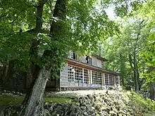Photo couleur d'une maison en bois à deux étages, entourée d'arbres au feuillage vert. La façade du rez-de-chaussée est presque entièrement fenêtrée.