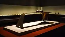 Photo couleur d'un brûle-parfum en bronze avec poignée, exposé au centre d'une salle de musée peu éclairée. En arrière-plan (flou et sombre), une vitrine éclairée forme un angle droit le long du mur de la salle.