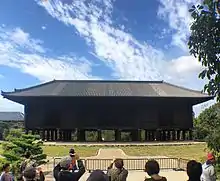 Photo couleur d'un large bâtiment en bois de couleur sombre. Un ciel bleu nuageux en arrière-plan, et, au premier plan, quelques personnes prenant des photos devant une barrière.