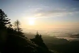 Photo couleur d'un lever de soleil au loin, au-dessus d'une couche de nuages, vu du sommet d'une montagne.
