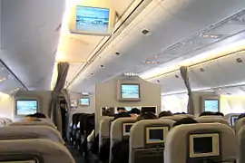 Cabne d'avion de ligne. Les rangée de sièges sont disposées autour de deux couloirs. Chaque dossier dispose d'un écran ; d'autres écrans sont accrochés au plafond.