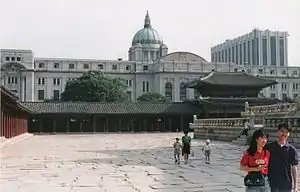Photographie prise depuis l'intérieur d'un ancien palais coréen. L'enceinte extérieure est le principal élément visible, avec plus loin à l'extérieur un grand bâtiment japonais qui domine l'ensemble.