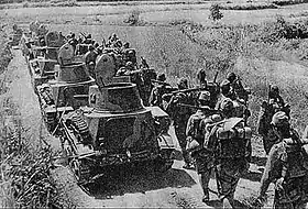 Photo en noir et blanc de la progression sur une route de chars de combat (à gauche) et de militaires (à droite).