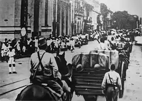 Photo ancienne représentant un convoi militaire, vu de dos, marchant dans une rue. On aperçoit des passants en arrière-plan.