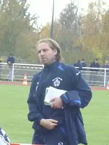 Photo d'un homme debout, vêtu d'un survêtement, présent sur un terrain d'entraînement de football et tenant un carnet dans sa main gauche.