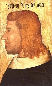 Portrait de profil d'un homme aux cheveux roux et bouclés, avec une légère barbe brune, portant un manteau noir simple au col blanc.
