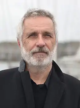 Portrait photographique d'un homme blanc d'une soixantaine d'années, cheveux et barbe courte blanche, veste et chemise noire.