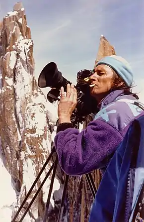 Un homme filme devant des montagnes, bonnet sur la tête et cigarette au bec.