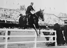 Photo en noir et blanc montrant un cavalier en uniforme franchissant une barrière blanche; les gradins étant combles derrière lui en arrière-plan.