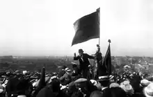 Photo noir et blanc d'un homme d'âge en chapeau melon tenant un drapeau uni et s'adressant à la foule.