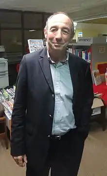 Un homme souriant, les traits vieillis, les cheveux clairsemés, la main gauche dans la poche du pantalon, est devant un stand dans une réception.