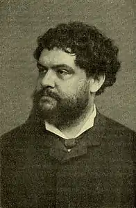 Photographie en noir et blanc d'un homme moustachu et barbu, aux cheveux bouclés.