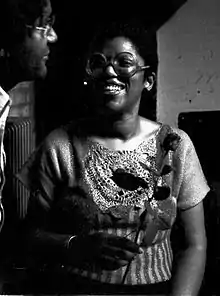 Photo en noir et blanc d'une femme noire souriante portant des lunettes rondes et tenant une rose à la main.