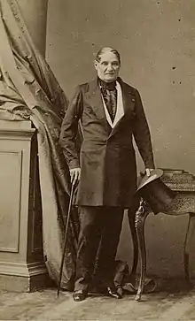 Photographie en noir et blanc de plein pied de Jérôme Bonaparte, âgé d'environ soixante-huit ans. Il porte un costume civil sombre, s'appuie sur une canne à gauche et tient un haut-de-forme à droite.