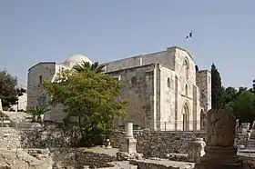 Vue générale d'une église en très bon état, entourée de verdure et de ruines antiques. Un drapeau français flotte au-dessus.