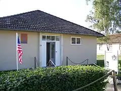 Un petit pavillon de plain-pied. Devant l'entrée, une haie et un drapeau américain. Un panneau annonce qu'il s'agit de la maison de Jesse Owens ; le reste du texte est illisible en raison de la luminosité trop haute.