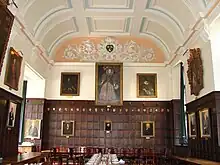 Un hall lambrissé de bois, avec un grand portrait d'Elizabeth I au milieu du mur du fond, d'autres  portraits sur les murs ; un plafond de plâtre blanc à décor bleu clair