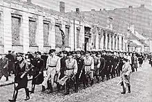 Photo en noir et blanc représentant plusieurs dizaines d’hommes coiffés de casquettes, matraque à la main, avançant en rangs de trois, dans une rue pavée.