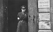 Un policier debout se tient dans l’embrasure d’une porte. À sa droite, une plaque sur le mur extérieur indique (en allemand puis en polonais) : « Service d’ordre du Judenrat de Varsovie, gestion du quatrième arrondissement ».