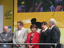 Photographie en couleur représentant cinq personnes, quatre de face et une de trois-quarts dos, derrière une rambarde et devant un panneau portant l'inscription : le tour de France 2014