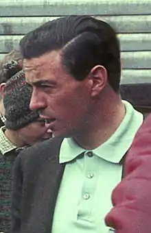 Photographie en couleur d'un homme brun, à la peau bronzée, vue de profil, en gros plan, avec un polo vert clair, et une veste marron.