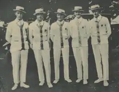 Photographie en noir et blanc montrant cinq hommes se tenant debout, en costume et pantalon blanc, portant chacun un canotier.