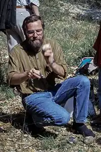 Photographie en couleur d'un homme à demi accroupi, portant une barbe et des lunettes et vêtu d'un jean et d'une chemise marron en train de reconstituer un débitage laminaire par percussion directe.