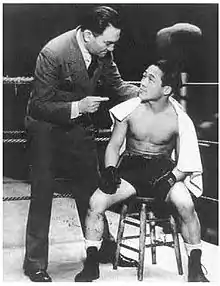 Photo noir et blanc d'un boxeur assis sur une chaise dans le coin d'un ring de boxe, torse nu, une serviette blanche sur le dos, fixant un homme en costume, debout à sa droite.