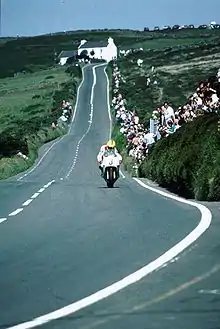 Photo du pilote Joey Dunlop en course, s'apprétant à négocier un virage après une longue ligne droite.