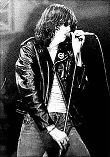 photo en noir et blanc d'un homme vêtu d'un jean et d'un blouson en cuir noirs chantant dans un micro.