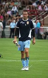 Photographie montrant Johann Durand arborant survêtement bleu et un maillot numéroté un qui tient dans sa main droite une paire de gant de gardien de but de football