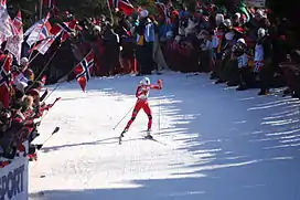 Vue lointaine d'une skieuse, avec de nombreux spectateurs de chaque côtés brandissant des drapeaux.