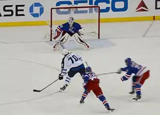 Photographie d'une partie de patinoire avec trois joueurs avec un uniforme de hockey bleu et un avec un uniforme blanc en train de tirer au but.