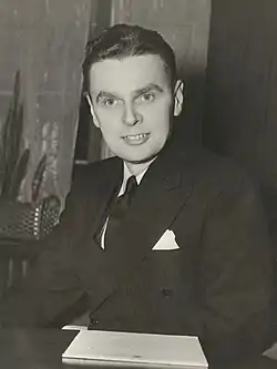 Un Diefenbaker souriant est assis derrière un bureau avec des documents de la Chambre des communes devant lui.