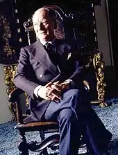 Vieil homme élégant et souriant, aux cheveux courts, vu de face, assis sur un fauteuil en bois, doigts et jambes croisées et observant sur sa droite.