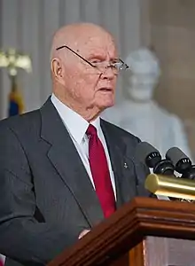 Un John Glenn plus âgé parlant sur un podium, avec ses lunettes perchées haut au-dessus de ses oreilles pour qu'il puisse lire avec elles