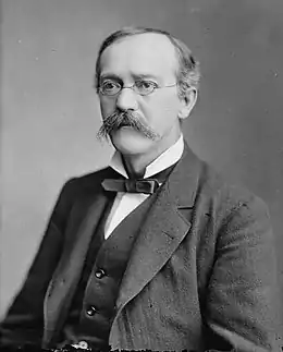 Un homme moustachu, portant des lunettes, en costume et nœud papillon.