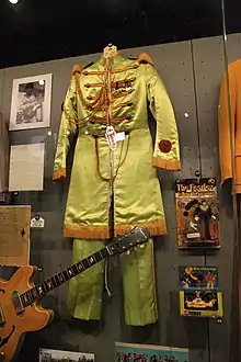 un uniforme de parade vert clair exposé dans une vitrine, guitare électrique au 1er plan