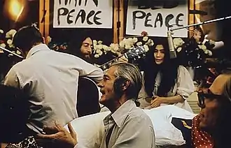 John Lennon (avec une guitare) et Yoko Ono, dans un lit entouré de gens dans une chambre d'hôtel.