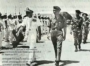 L'amiral, en uniforme blanc, passe en revue une troupe ; Sirik Matak est en uniforme sombre à sa gauche, accompagné de deux officiers en retrait.