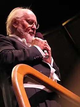 Un homme à la barbe et aux cheveux blancs, portant un costume noir, se tient sur une estrade et parle en tenant un micro dans ses mains.