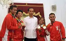 Un homme en chemise blanche se tient debout entre quatre athlètes en survêtements rouges qui brandissent des médailles.
