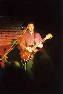 Chante sur scène, en chemise rose, jouant de la guitare semi-acoustique.