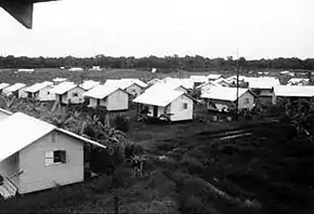 Photo en noir et blanc montrant de nombreuses petites maisons blanches identiques sur un terrain plat en terre.