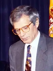 Photographie d'un homme aux cheveux gris, portant des lunettes. Il est assis et regarde vers le bas.