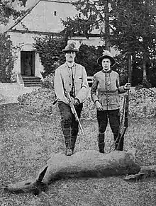 Posant devant une dépouille de gibier, deux jeunes hommes portant une tenue de chasse et portant un fusil.