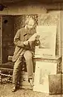 Homme barbu en blouse, debout près d'un chevalet, penché sur un document