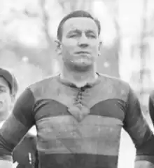 Photographie noir et blanc : c'est un portait en buste d'un homme en maillot de sport qui regarde devant lui. À gauche de l'image est visible la moitié d'un visage. L'arrière-plan est flou et on devine des arbres.