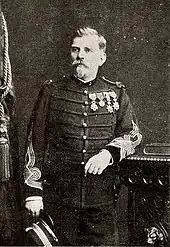 Photographie noir et blanc d'un homme debout en uniforme, le coude sur un meuble.