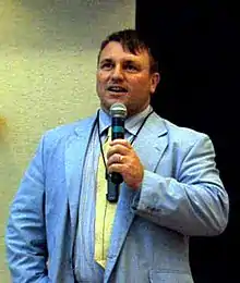 Portrait en buste de Joseph Pearce, chemise rayée bleue et veste de costume bleue, cravate jaune, tient un micro dans sa main gauche.